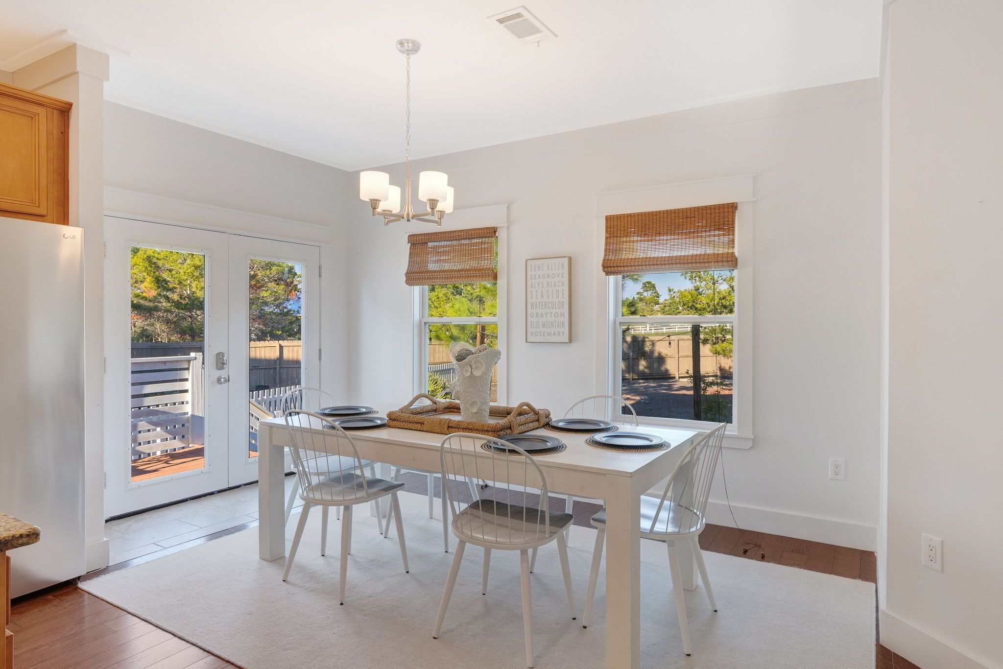 A dining area with a white table set for four, surrounded by white chairs, a chandelier, and two windows with blinds.