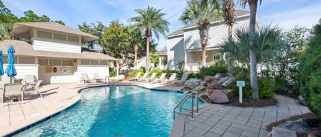 A swimming pool with lounge chairs and palm trees, surrounded by buildings with a white exterior.