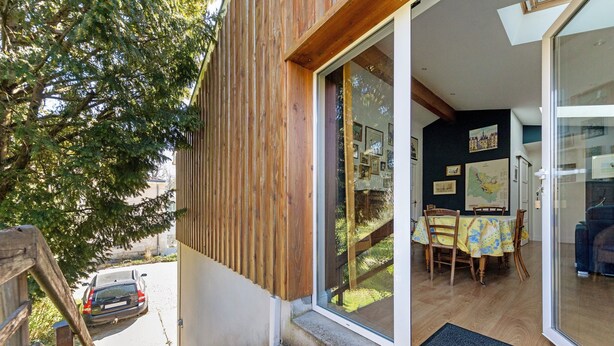 Un mur extérieur en bois, une porte vitrée donnant sur un espace de repas intérieur avec une table et des chaises, et une voiture garée à l’extérieur.