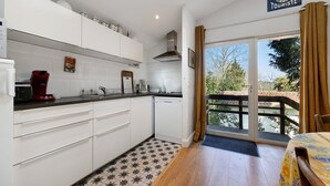 Une cuisine avec des meubles blancs, une machine à café rouge et une vue sur un balcon doté d’une balustrade en bois.