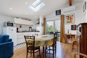 Una cocina moderna con armarios blancos, una mesa de comedor con sillas, un sillón azul y vistas a través de una ventana.