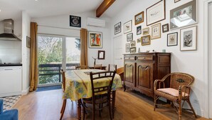 Un coin repas avec une table, des chaises et un fauteuil en osier. Des tableaux encadrés ornent le mur, et une enseigne est accrochée au-dessus de la table.