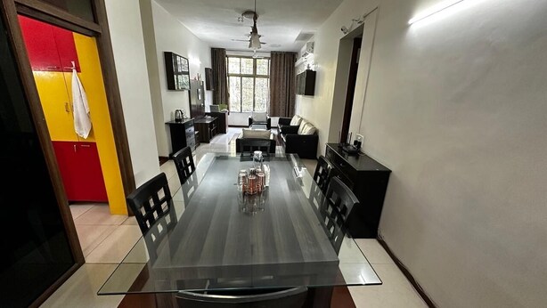 A dining area with a glass-top table, black chairs, a red and yellow cabinet, and a kitchen in the background. - Amicable Corporate Apartment (Mumbai)