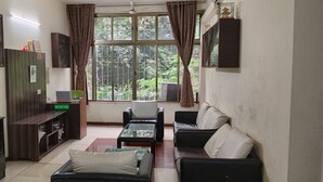 Un salon comprenant un canapé, une table basse, un bureau avec un ordinateur et un meuble. Il y a une fenêtre avec des rideaux, et de là, on peut admirer la verdure à l’extérieur.
