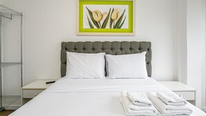 Un letto rifatto con lenzuola bianche e due asciugamani piegati, una testiera grigia trapuntata e un quadro con fiori appeso al muro.