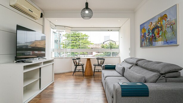 Un soggiorno moderno con un divano grigio, una TV su un mobile bianco, un tavolo da pranzo con sedie e un'ampia finestra vista alberi e palazzi.