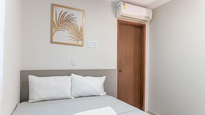 Une chambre à coucher avec un lit, des oreillers blancs, une œuvre d’art encadrée, un appareil de climatisation et une porte en bois.