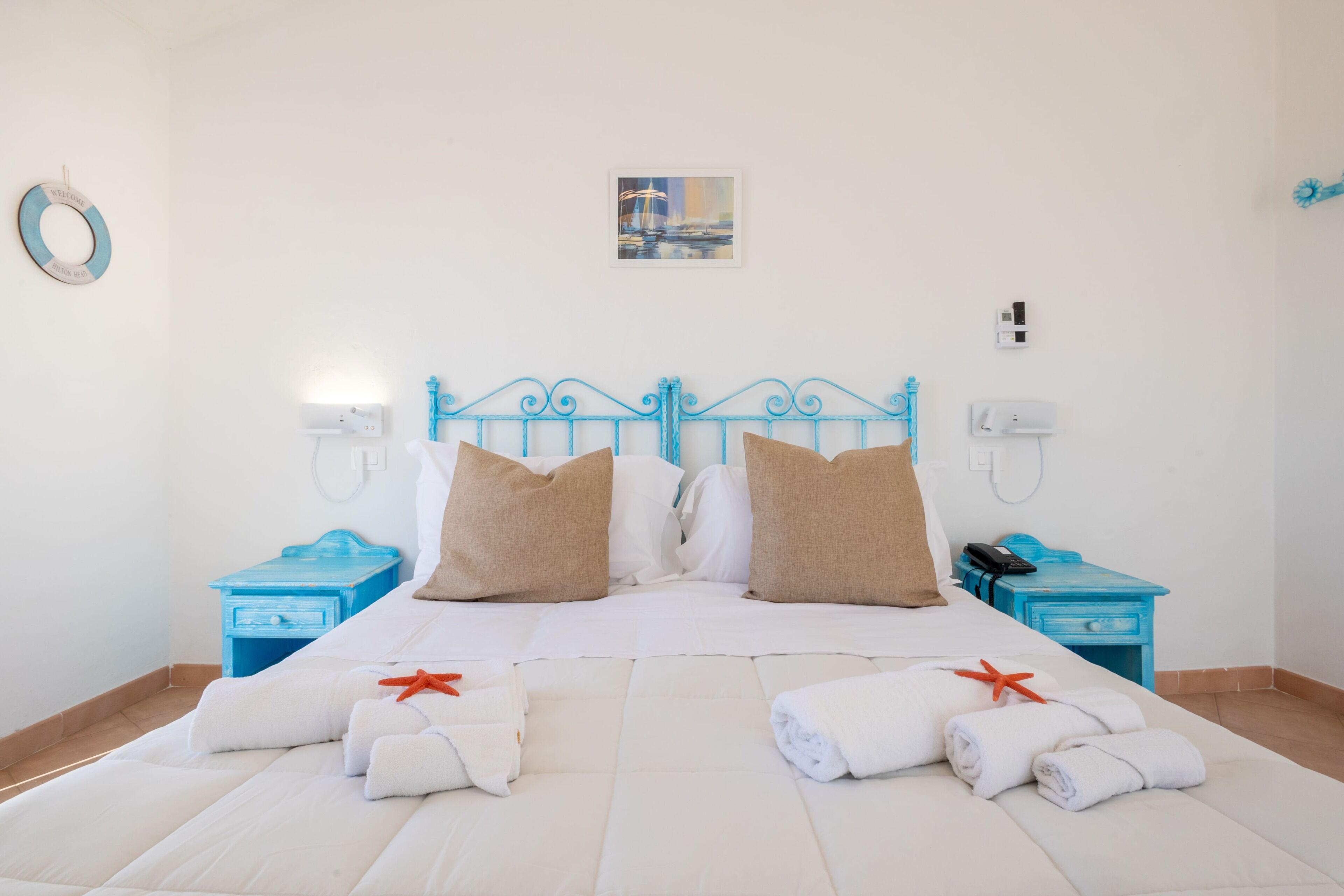 Una cama bien hecha con sábanas blancas y dos almohadas, un cabecero azul y un pequeño cuadro en la pared.