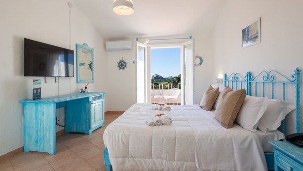 A bedroom with a bed, a desk, a television, and a balcony with a view. - Hotiday Baja Sardinia Seaside (Baja Sardinia)