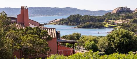 A coastal building with a red facade, a chimney, and a view of the sea and hills.