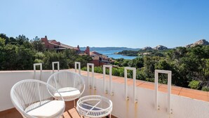 A terrace with white wicker chairs and a glass table, overlooking a scenic landscape with hills and a body of water. - Hotiday Baja Sardinia Seaside (Baja Sardinia)