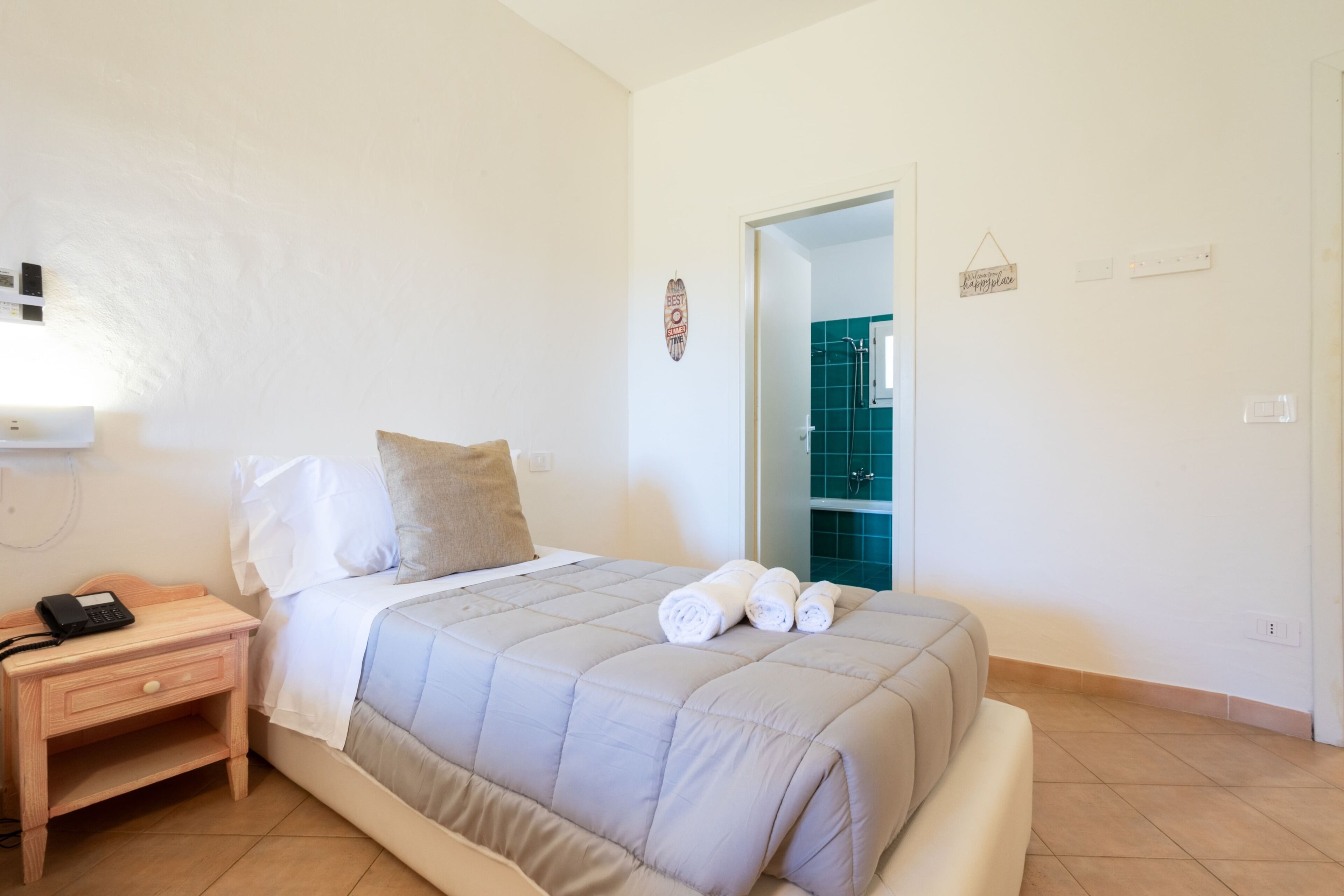 A bedroom with a bed, a nightstand with a phone, and a bathroom visible through an open door.