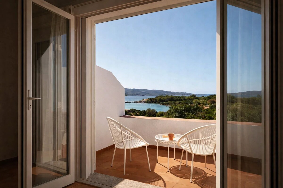 Un balcón con mesa y sillas, una puerta de cristal y vistas a un paisaje con agua y colinas.