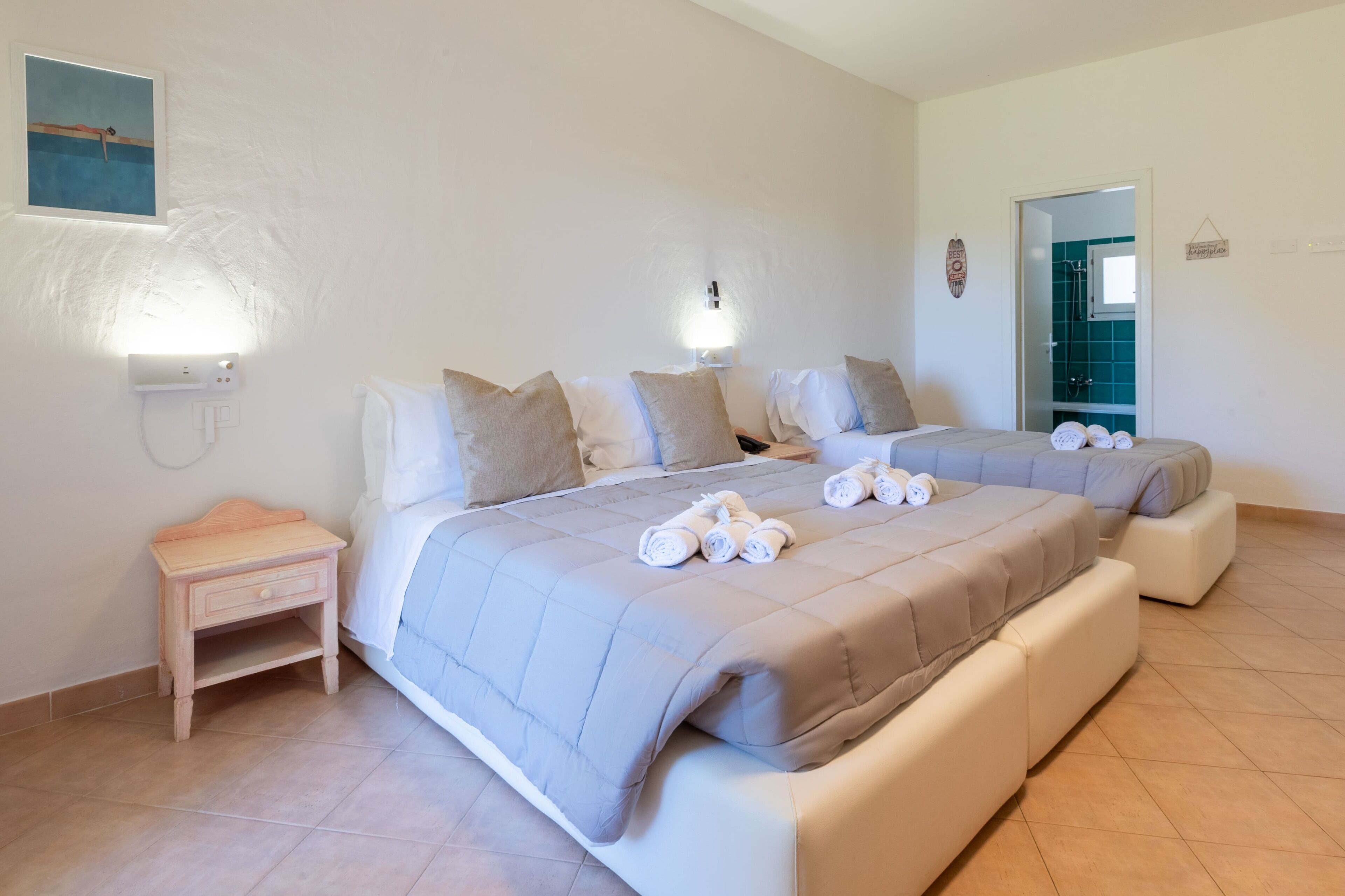 A bedroom with two beds, a nightstand, wall-mounted lights, and a door leading to a bathroom.