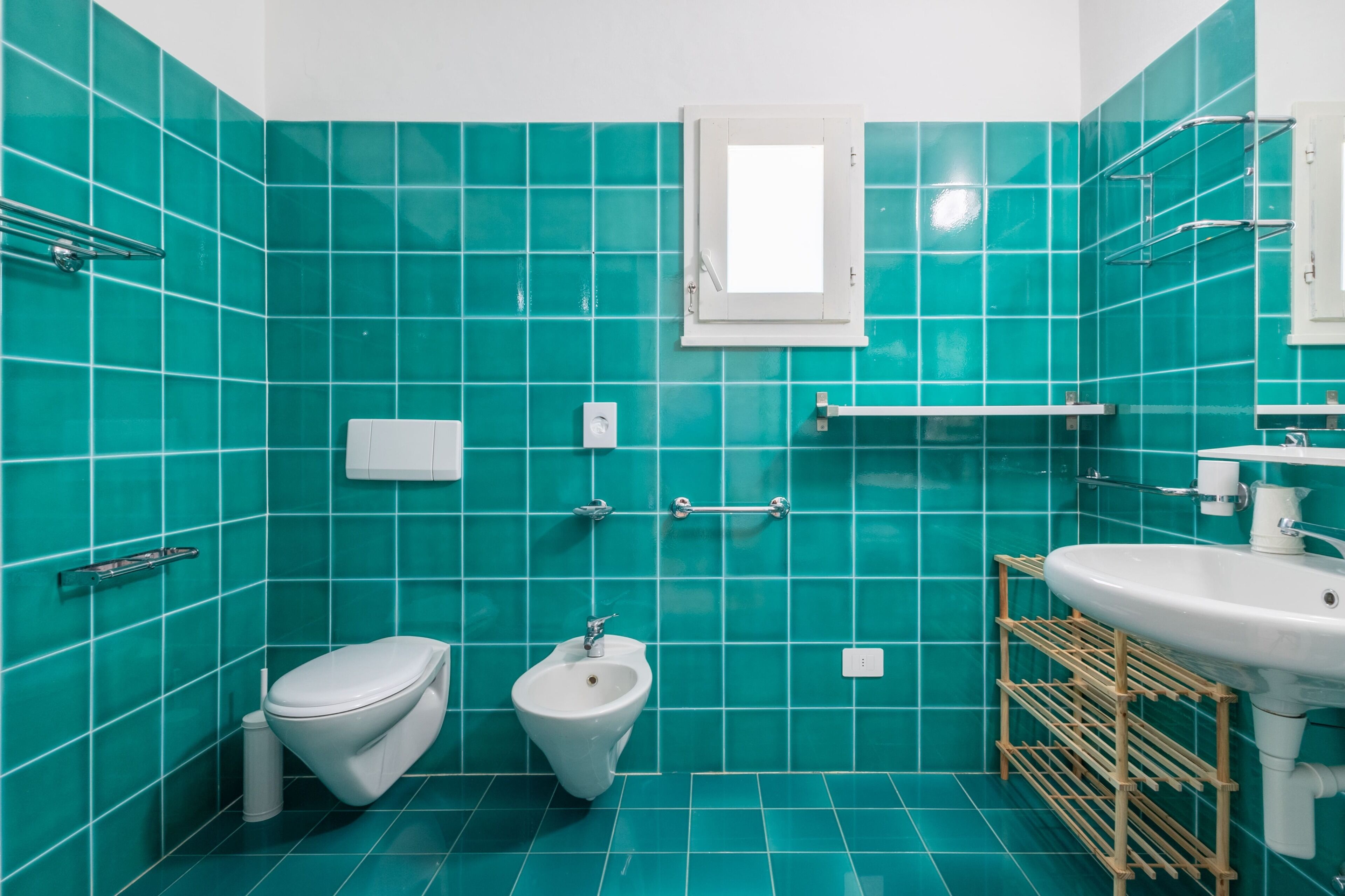 A bathroom with teal tiles, a toilet, bidet, sink, and a window.