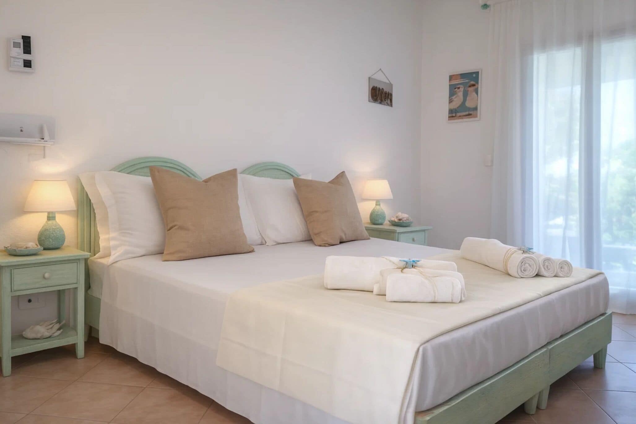 A neatly made bed with pillows and towels, a bedside lamp, and a small shelf with a vase.