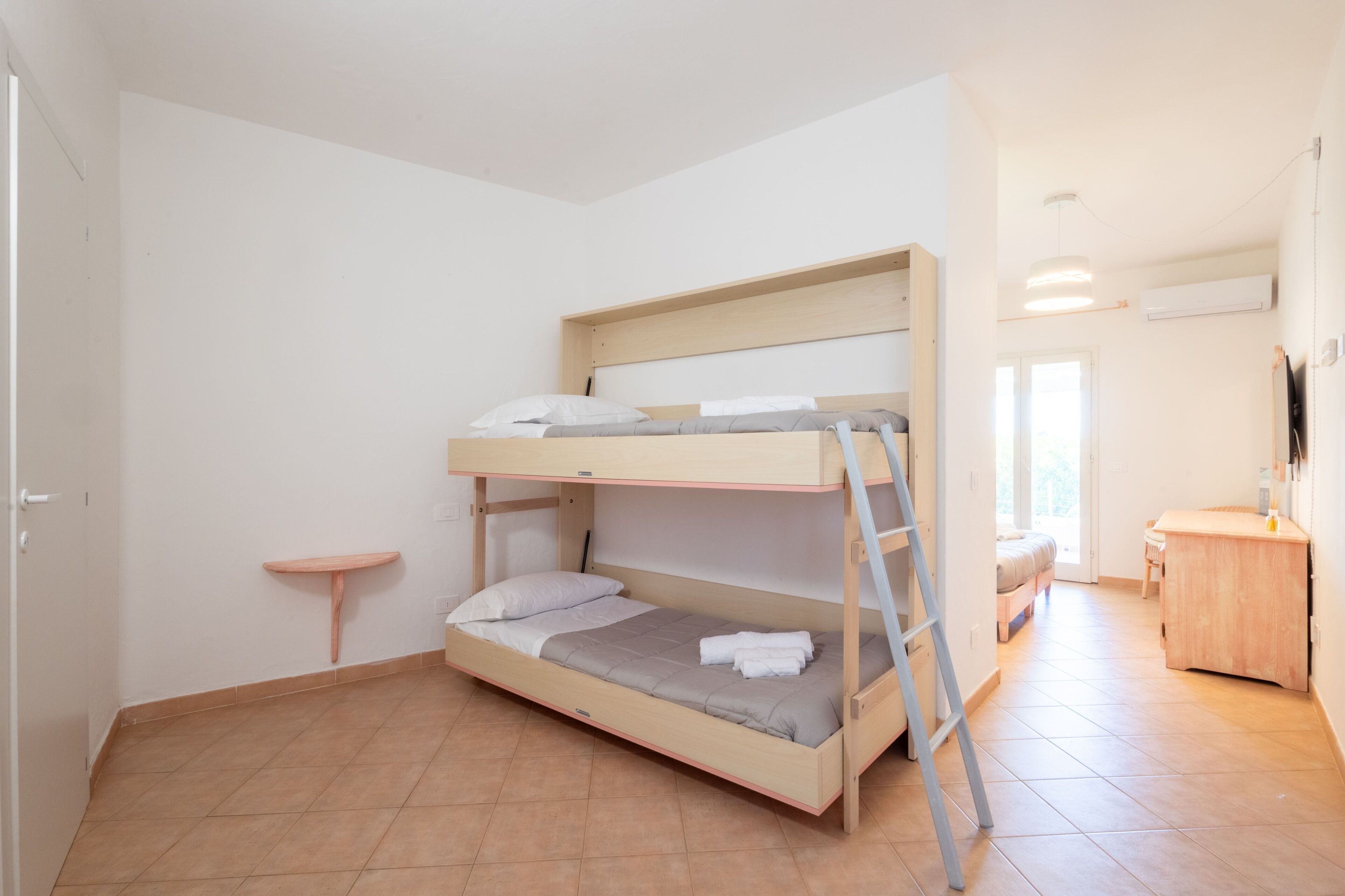 Una cama individual con escalera, un estante pequeño, un escritorio y una silla en una habitación con suelo de baldosas y paredes blancas.