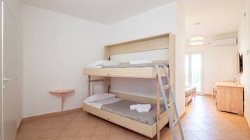Una cama individual con escalera, un estante pequeño, un escritorio y una silla en una habitación con suelo de baldosas y paredes blancas.