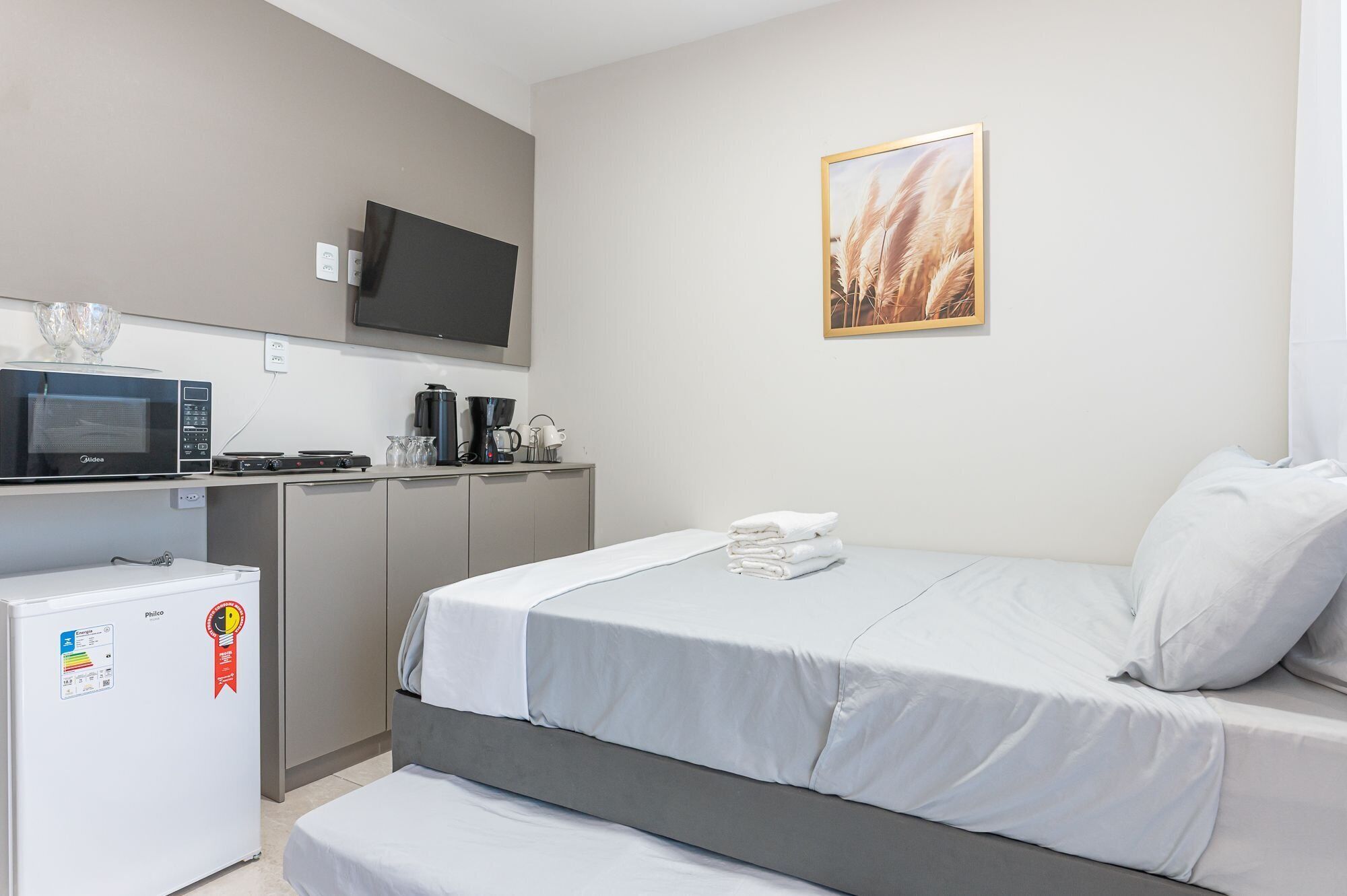 A compact hotel room with a bed, a small kitchenette, a television, and a framed picture on the wall.