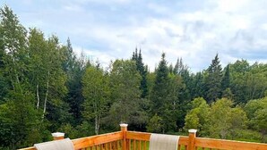 Une table à plateau de verre, accompagnée de quatre chaises, est placée sur une terrasse en béton, entourée d’une rambarde en bois et offrant une vue sur une zone boisée.