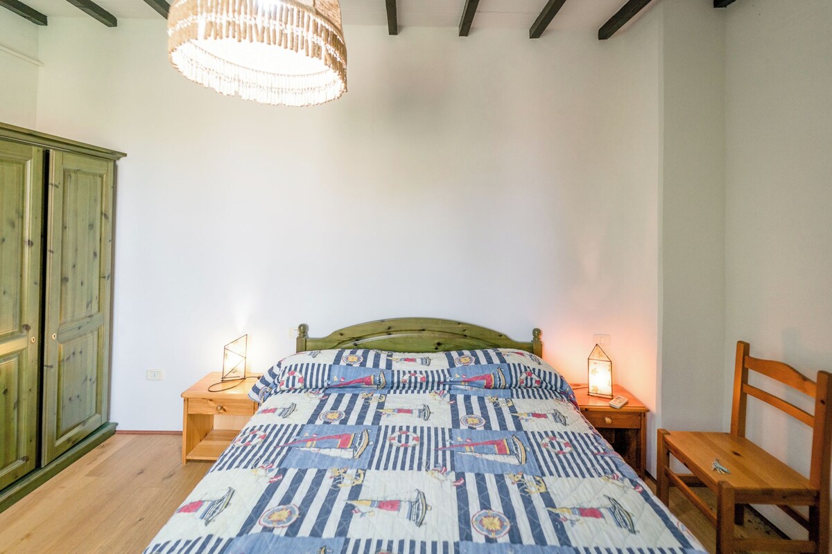 A bedroom with a bed, two bedside tables, a nightstand, a wooden chair, a green wardrobe, and a hanging light fixture.