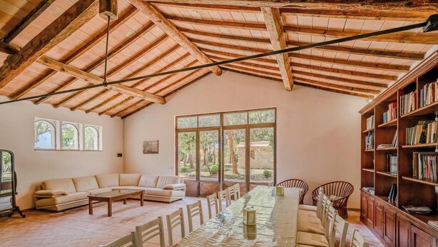 A spacious room with a high wooden ceiling, a large window, a sofa, a dining table, and a bookshelf. - Hotiday Grosseto Podere (Grosseto)