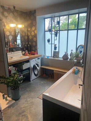 Une salle de bain moderne équipée d’un lave-linge, d’un évier et d’une baignoire. Une fenêtre offre une vue sur les arbres, et une étagère est décorée de plantes.