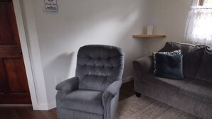 Een woonkamer met een grijze fauteuil, een grijze bank, een tapijt met een patroon, een houten deur en een wandplank met een wit object.