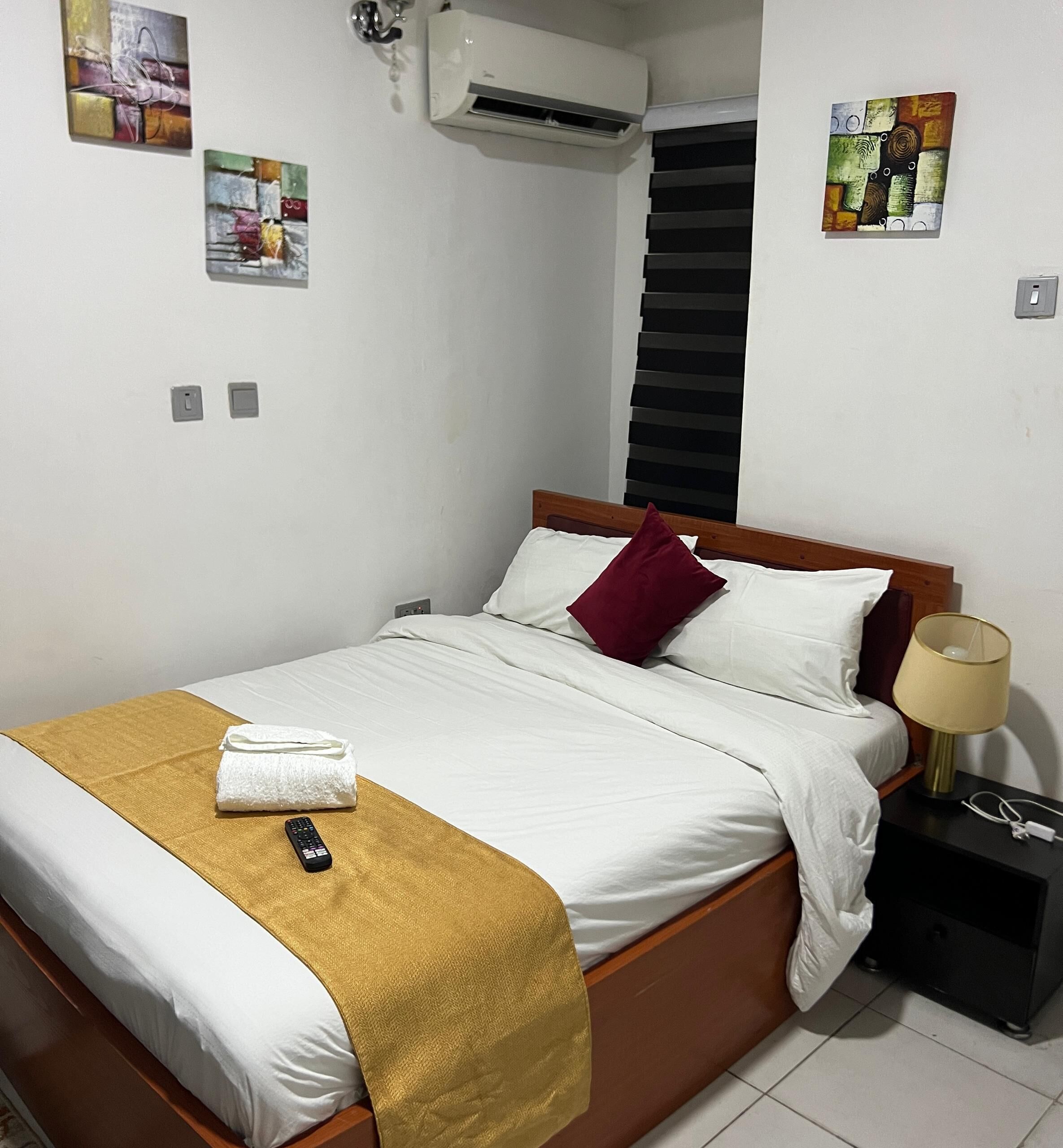 A neatly made bed with a gold throw blanket, a remote control, and a white towel. There is a bedside table with a lamp and a power outlet. The room has white walls and artwork on the walls.
