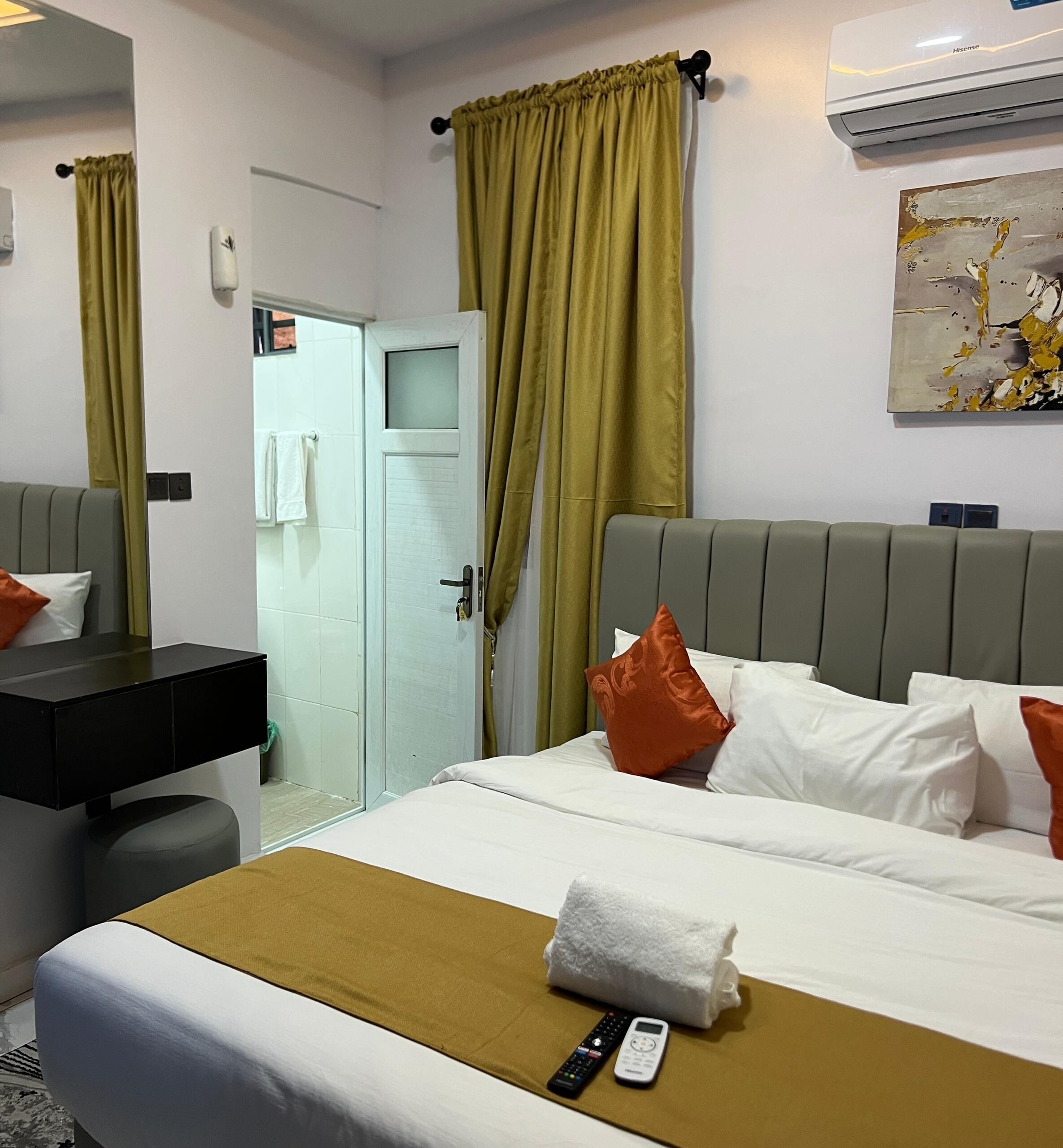 A hotel room with a bed, a bedside table, a mirror, a door, and an air conditioning unit.