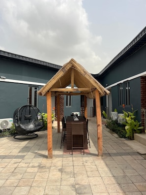 A covered outdoor seating area with a table and chairs, a hanging chair, and a thatched roof structure. - RBZ Apartments (Lagos)