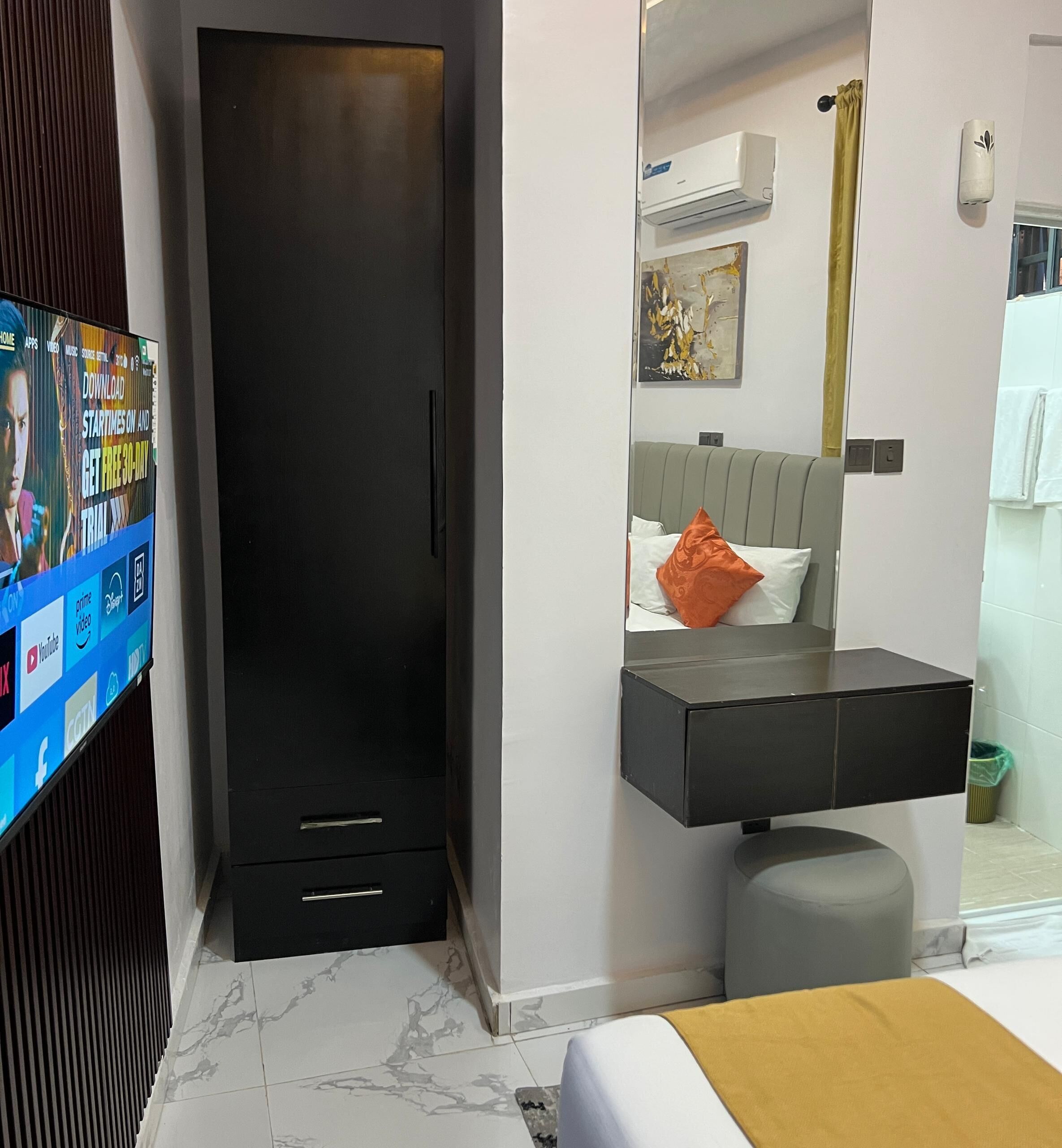 A modern hotel room with a black wardrobe, a grey ottoman, a bed with a yellow throw, a grey headboard, and a small bathroom with a toilet and sink.