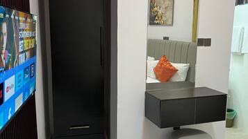 A modern hotel room with a black wardrobe, a grey ottoman, a bed with a yellow throw, a grey headboard, and a small bathroom with a toilet and sink.
