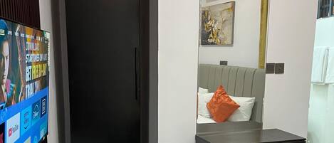 A modern hotel room with a black wardrobe, a grey ottoman, a bed with a yellow throw, a grey headboard, and a small bathroom with a toilet and sink.