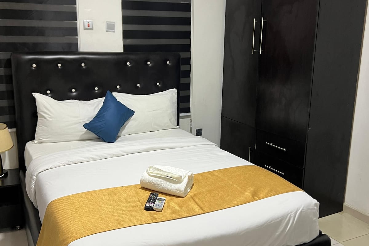 A neatly made bed with white and gold bedding, a blue pillow, and a folded towel on a gold throw. A remote control is placed on the bed.