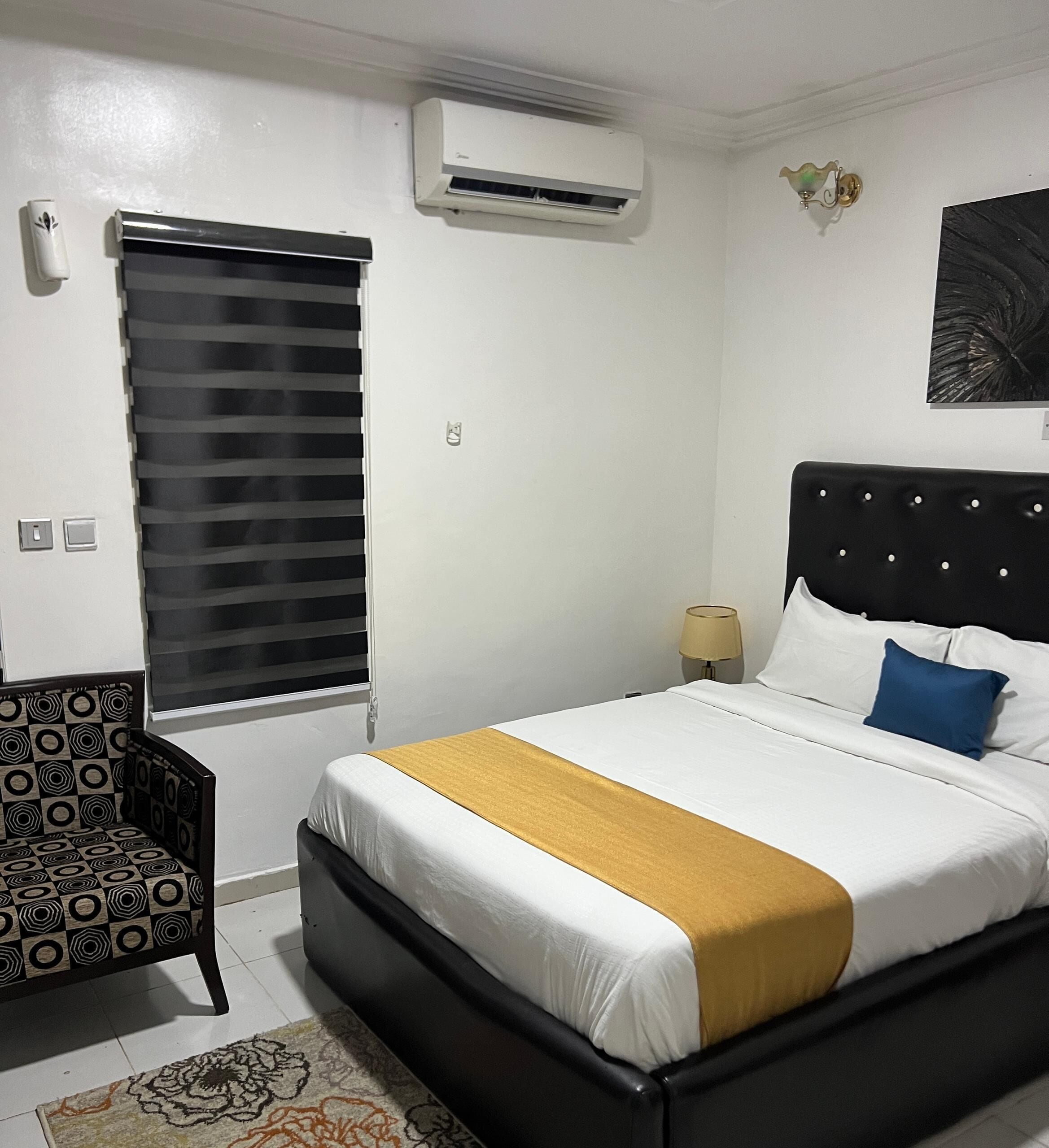 A hotel room with a bed, a chair, an air conditioner, a lamp, and a painting on the wall.