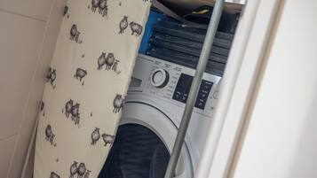 A laundry area with a washing machine, a shelf with books, and a patterned curtain.