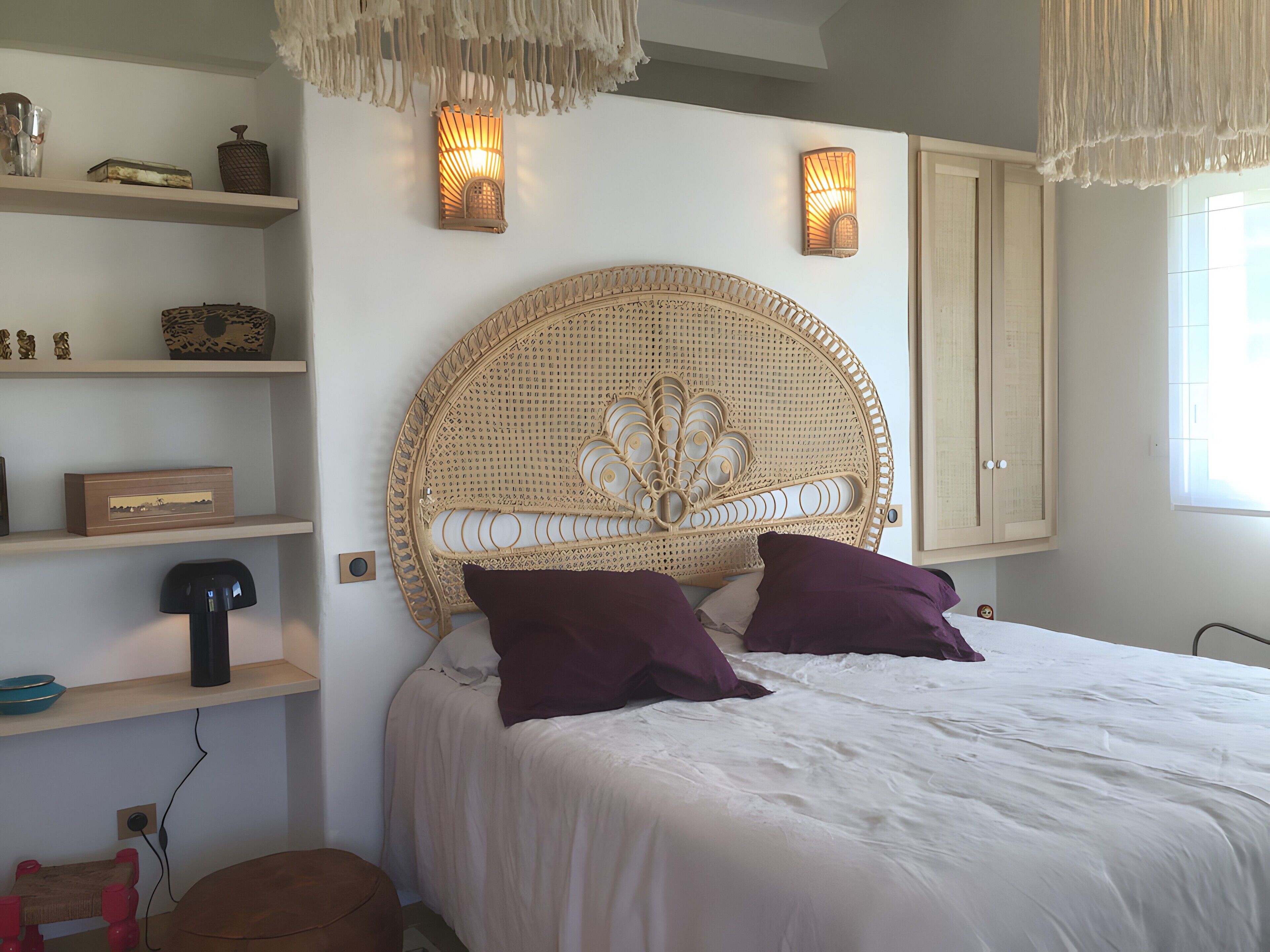 Une chambre à coucher avec une tête de lit en rotin, deux lampes fixées au mur, un lit avec une literie blanche et une fenêtre laissant entrer la lumière naturelle.