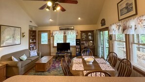 A living room with a sofa, TV, dining table, and ceiling fan. - OC80: Suite: Canal front with private boat dock and large yard (Ocracoke)