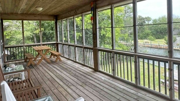 A covered deck with wooden flooring, a picnic table, and a railing overlooking a body of water. - OC80: Suite: Canal front with private boat dock and large yard (Ocracoke)
