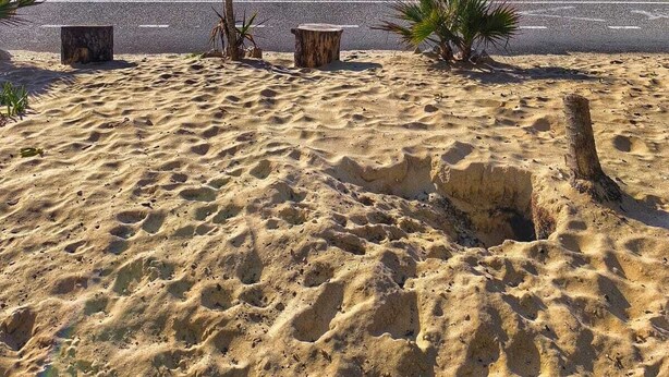 Una playa con un paseo pavimentado, palmeras y un promontorio rocoso a lo lejos.
