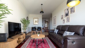 Ein modernes Wohnzimmer mit einer Ledersofa, einem Fernseher, einem Essbereich mit Stühlen und einem Tisch, und einer großen Pflanze.