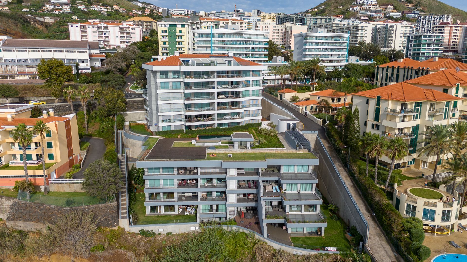 Un hôtel moderne à plusieurs étages, doté d’une piscine sur le toit, entouré d’autres bâtiments et de verdure.