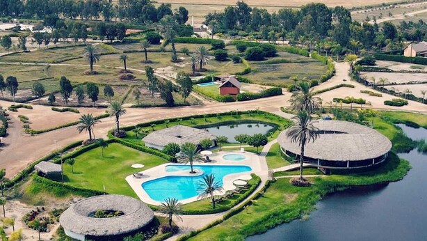 An aerial view of a landscaped property with a central swimming pool, circular structures, and a surrounding road.