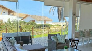 A modern living room with wicker furniture, large windows, and a view of a beach and buildings.