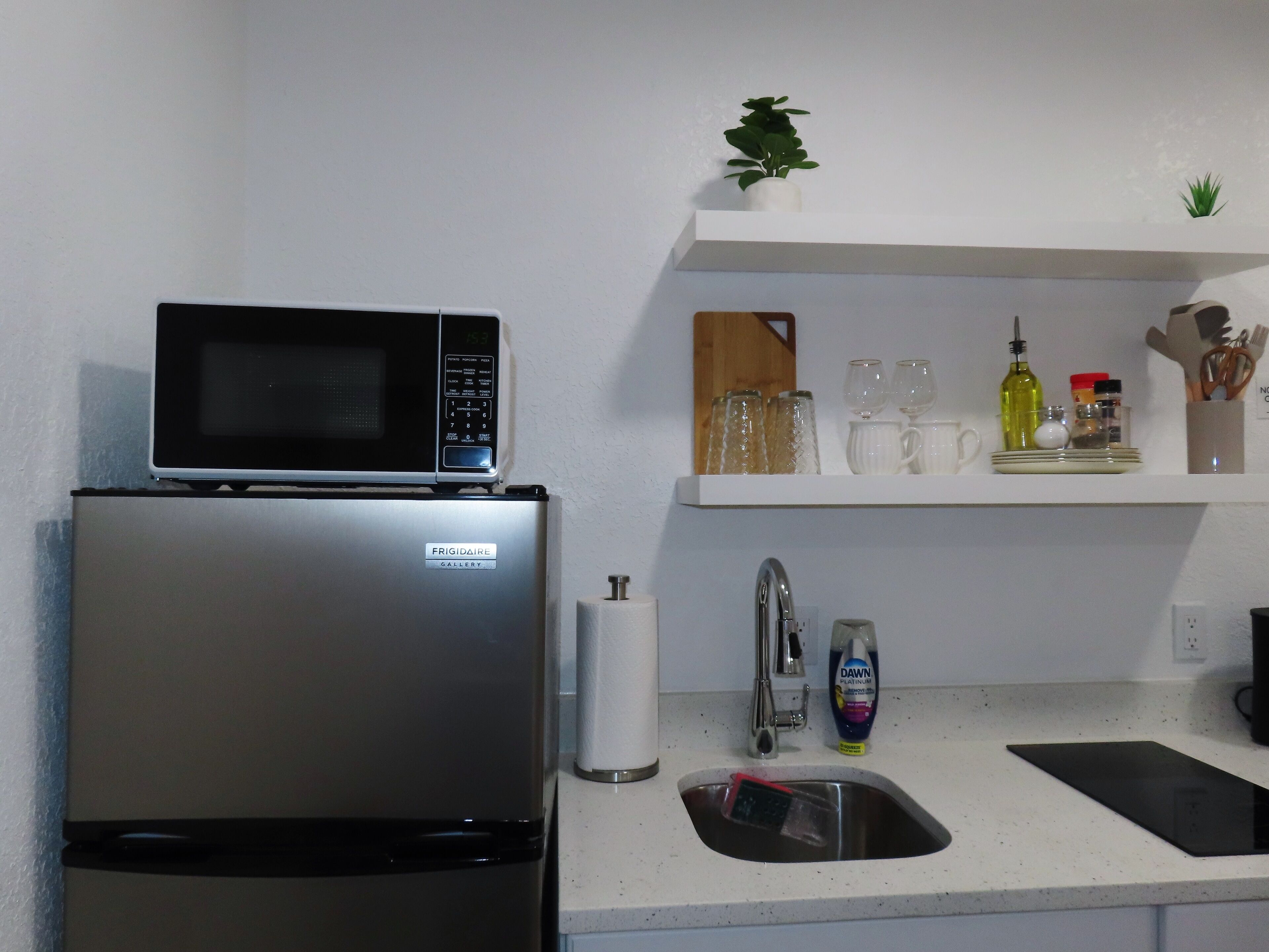 Una cocina con un refrigerador de acero inoxidable, un microondas, un lavamanos y una repisa sobre la encimera con varios utensilios de cocina.