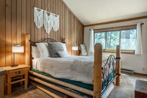 A bedroom with a wooden bed frame, a nightstand with a lamp, a macrame wall hanging, and a window with a view of trees. - Shared Hot Tub and Pool | Mountain Views (Mammoth Lakes)