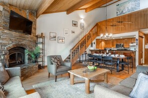 A spacious living area with a fireplace, a dining table with chairs, a kitchen with wooden cabinets, and a staircase leading to an upper level. - Shared Hot Tub and Pool | Mountain Views (Mammoth Lakes)