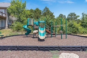 Ein Spielplatz mit Rutsche und Klettergerüsten, umgeben von Bäumen und einem Gebäude im Hintergrund.