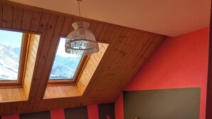 Ein Schlafzimmer mit schräg steilender Decke, Holzbalken, einem Bett mit weißer Bettwäsche, einem Nachttisch mit Lampe und Blick auf schneebedeckte Berge durch Dachfenster.